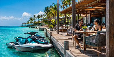 A jet ski next to a water front restaurant dock with a male and female sitting at a dining table.